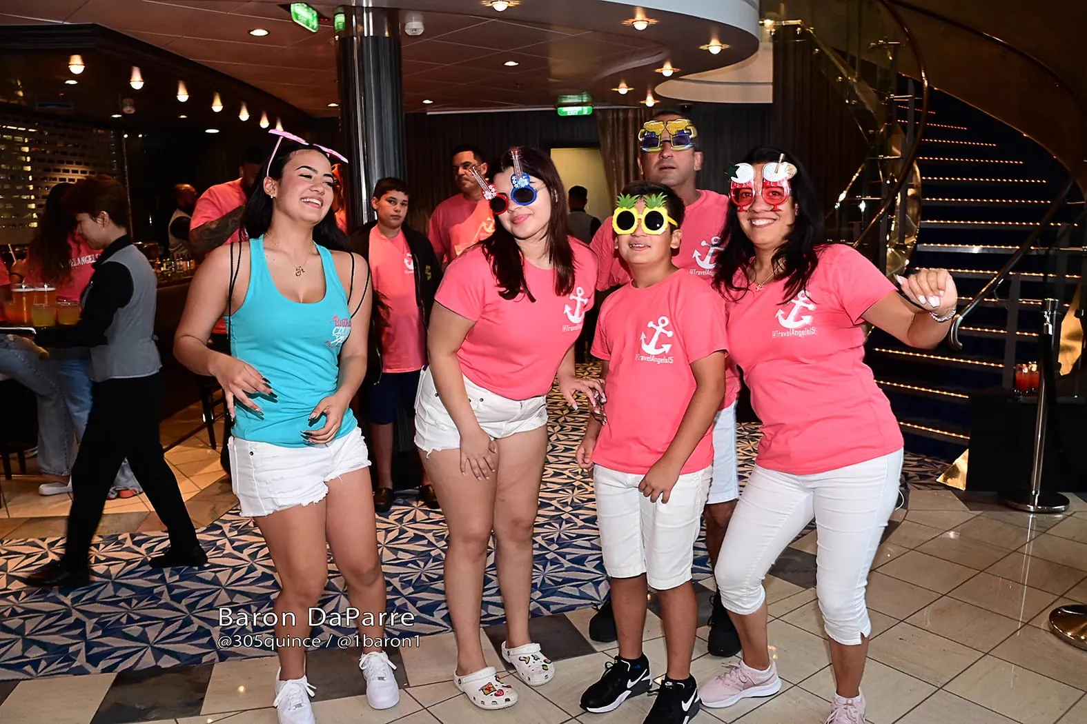 A group gathers indoors wearing pink Travel Angels shirts and festive glasses.