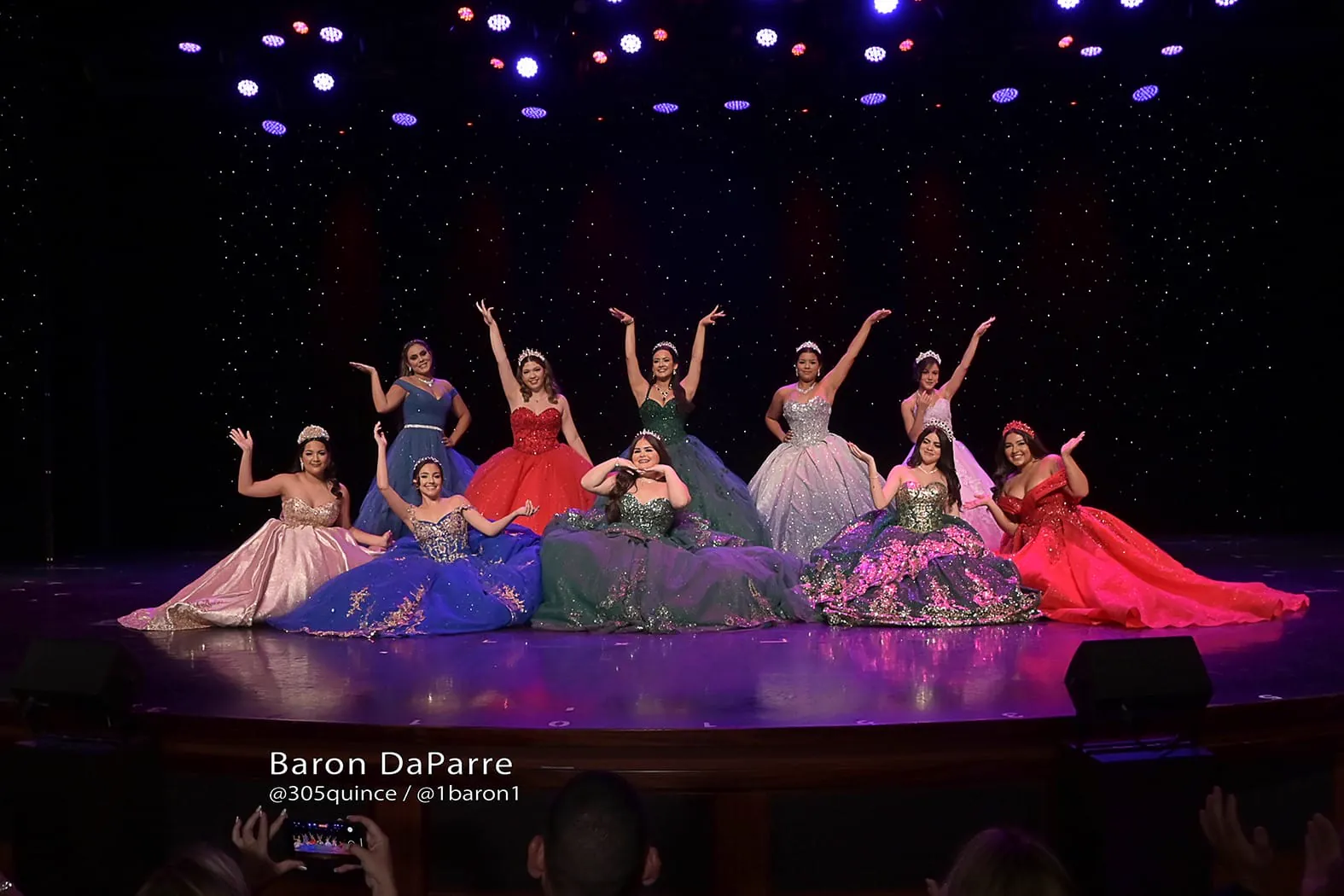 A group in formal gowns poses on a stage under bright lights.